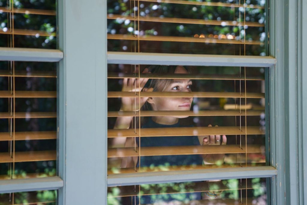 Young woman looks in big window with white frame through protective brown shutter enjoying view of summer nature in yard. Lush green leaves of trees reflected on clean window glass surface close view