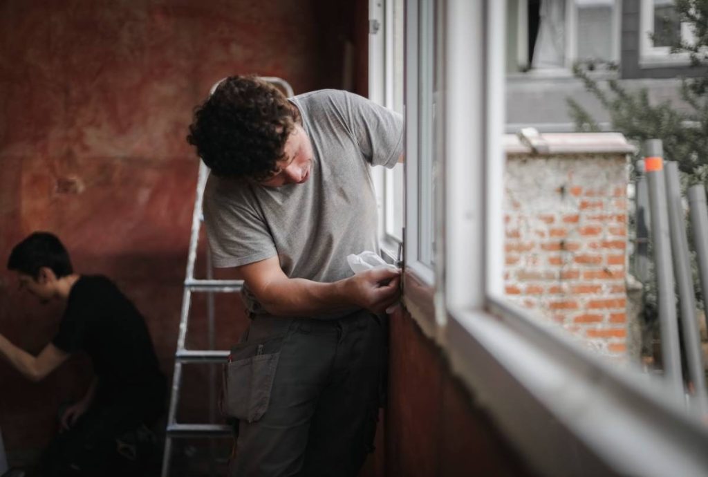 Two caucasian men in uniforms wash window frames in a room with red walls where repairs are underway, close-up side view with selective focus. The concept of home renovation, washing window frames.